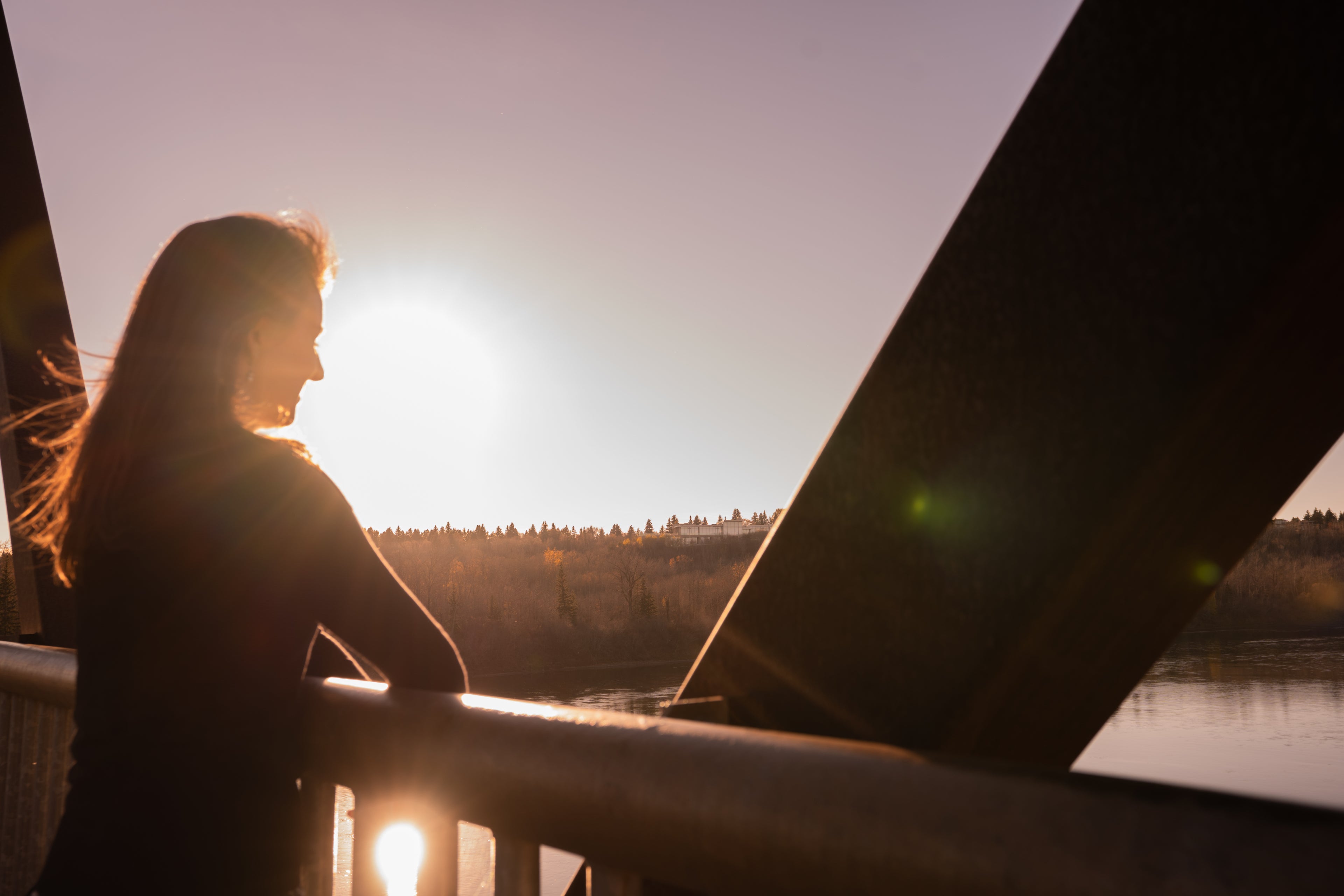 Woman standing on a bridge over a river at sunset, silhouetted against the sun.